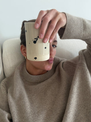 Young woman holding a beige ceramics cup wiht lupine coffee with black splashes on it, wearing a beige cashmere sweater, sitting on a beige couch. 