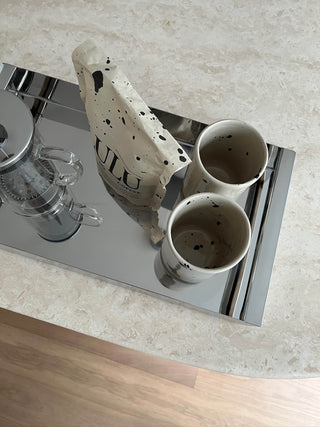 Lulu The No Coffee Lupinenkaffee Verpackung auf einem modernen Edelstahltablett zusammen mit einer French Press aus Glas und zwei Keramikbechern in Beige mit schwarzen Punkten auf einem Travertintisch, Perspektive von oben.