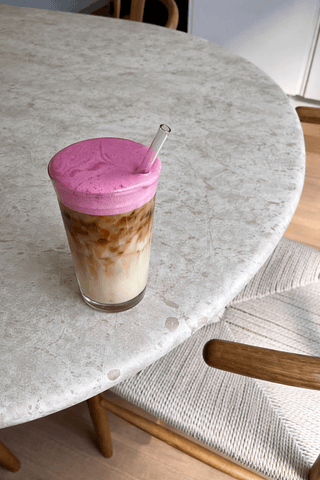 Lulu Blush: A tall glass filled with iced lupine coffee, topped with a generous cloud of soft pink whipped cream, garnished with a pinch of cardamom and a glass straw. Natural light, minimal background.