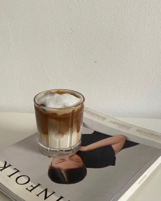 A layered lupine drink in a clear glass with foamed milk on top, sitting on a fashion magazine. The glass is placed over a model's face on the cover.