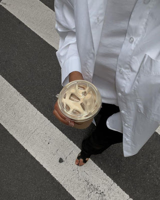 Person in a white button-up shirt holding a clear plastic cup of cold Lulu lupine coffee, standing on a gray pavement with white stripe markings.