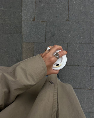 A hand holding a white to-go cup lid, wearing silver rings, sleeve of a beige trench coat visible. Shot from above against a grey stone pavement.