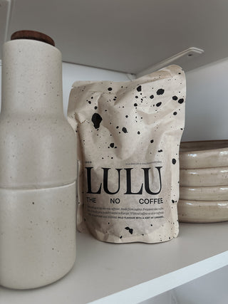 Lulu-Verpackungsbeutel mit charakteristischem Tintenspritzer-Muster auf einem Regal, neben gestapelten Objekten.