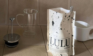 A bag of Lulu The No Coffee standing on a tiled countertop next to a glass French press carafe and a white ceramic cup. The packaging is cream-colored with black ink splatter details and bold black typography.