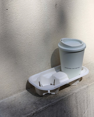 Reusable Coffee Cup in beige with the slogan "mornings, but better" standing on stairs outside in front of a beige wall, play of light and shadow.
