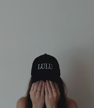 A young woman wearing a black hat with the slogan "lulu" hiding behind her hands with golden rings in front of a white wall.