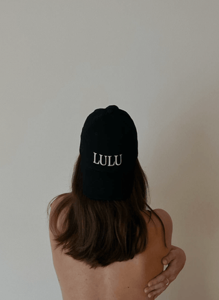 Young woman with brown hair wearing a black cotton cap the other way around with the logo "Lulu" on it, facing a white wall.