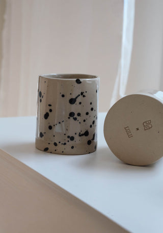 Beige Handmade Ceramic Cup with black splashes standing on a white table in front of a white curtain, another cup showing the buttom with the logos "lulu" and "studio babo".