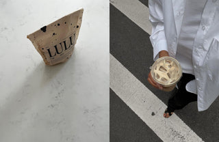 Person in a white button-up shirt holding a clear plastic cup of cold Lulu lupine coffee, standing on a gray pavement with white stripe markings.