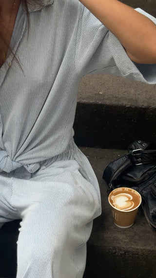 Woman sitting on stairs in a summer outfit one suite having a lupine coffee in a to go cup next to her a black leather purse in ninety style in an urban environment.