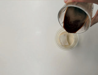 On a white backgorund a hand is pouring lupine coffee in a glass filled with iced cubes and oat milk.