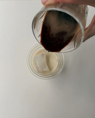 On a white backgorund a hand is pouring lupine coffee in a glass filled with iced cubes and oat milk.